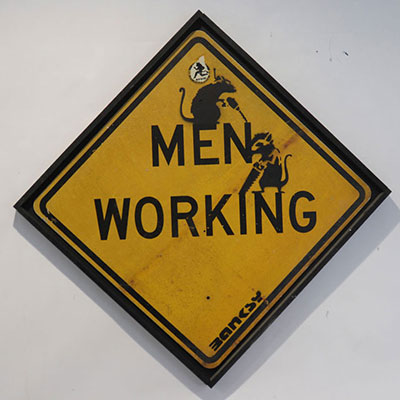 Banksy. “Men Working”. Black spray on yellow road sign.